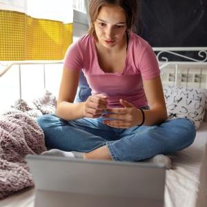 A young girl has psychotherapy session with her therapist via video call. She is sitting on her bed and talking with her therapist.